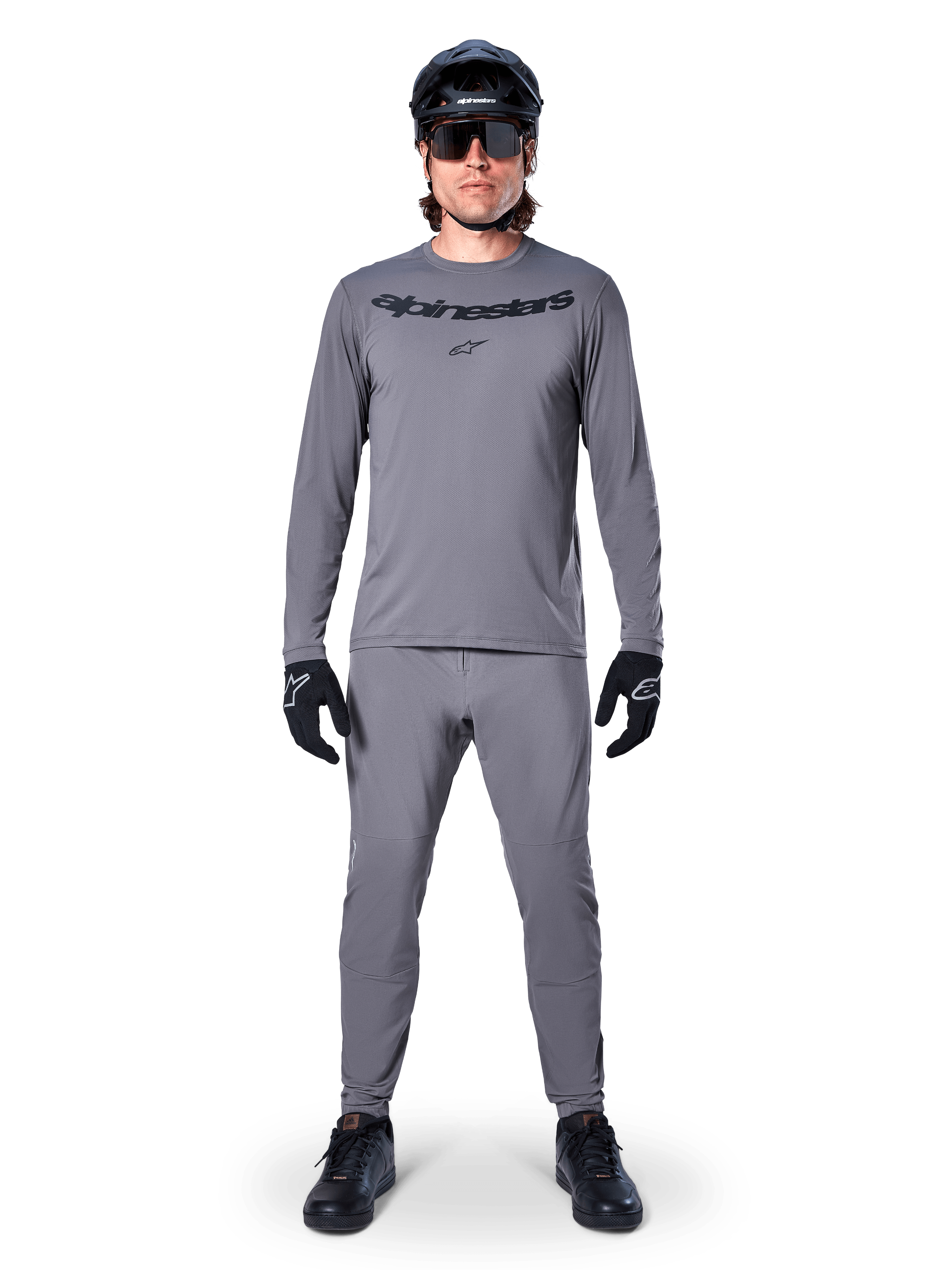 Mannelijke fietser in Alpinestars A-Dura Rocker Jersey met lange mouwen in donkergrijs, bijpassende grijze broek, zwarte MTB-helm, zonnebril en handschoenen, professionele mountainbikekleding voor off-road paden