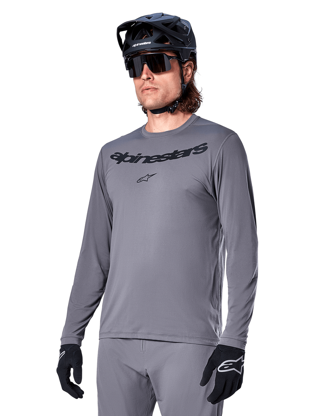 Alpinestars A-Dura Rocker jersey met lange mouwen, grijs MTB-fietsshirt, gedragen met zwarte helm en handschoenen, voorzien van zwart woordmerk op de borst, sportieve fietskleding voor off-road ritten, egale achtergrond