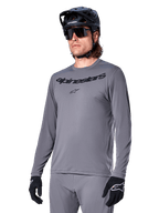 Alpinestars A-Dura Rocker jersey met lange mouwen, grijs MTB-fietsshirt, gedragen met zwarte helm en handschoenen, voorzien van zwart woordmerk op de borst, sportieve fietskleding voor off-road ritten, egale achtergrond