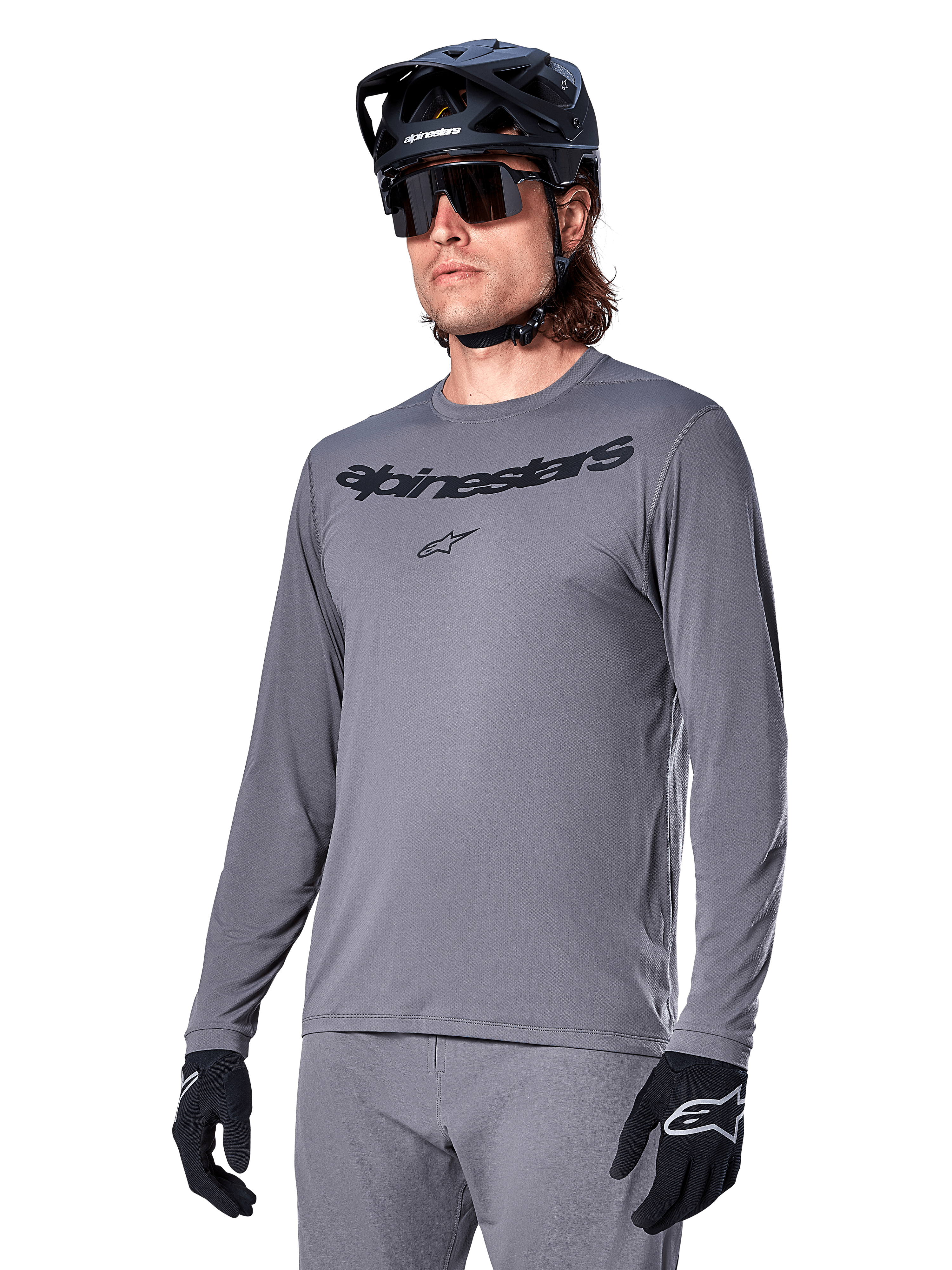 Alpinestars A-Dura Rocker jersey met lange mouwen, grijs MTB-fietsshirt, gedragen met zwarte helm en handschoenen, voorzien van zwart woordmerk op de borst, sportieve fietskleding voor off-road ritten, egale achtergrond