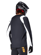 Cycliste de VTT portant un maillot à manches longues Alpinestars A-Supra Oscar, noir avec des empiècements blancs sur les épaules et les manches, vue de dos, casque et gants noirs, pantalon avec des touches d'orange, équipement de cyclisme tout-terrain durable