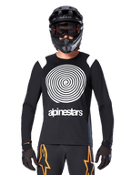 Maillot à manches longues Alpinestars A-Supra Oscar, noir avec graphique en spirale blanc et accents aux épaules, porté par un cycliste avec un casque intégral noir et des lunettes, pantalon noir et orange, équipement de cyclisme de performance pour le VTT gravity