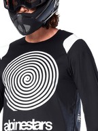 Maillot Alpinestars A-Supra Oscar, manches longues, noir, avec un grand graphique en spirale Oscar blanc sur la poitrine et le logo de la marque, porté par un pilote avec un casque intégral noir et un masque, vêtement de VTT haute performance pour la pratique du gravity