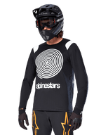 Alpinestars A-Supra Oscar Jersey à manches longues, maillot de VTT, noir et blanc, avec un motif en spirale blanc et le logo Alpinestars sur la poitrine, porté avec un casque intégral et un masque noirs, équipement de VTT haute performance