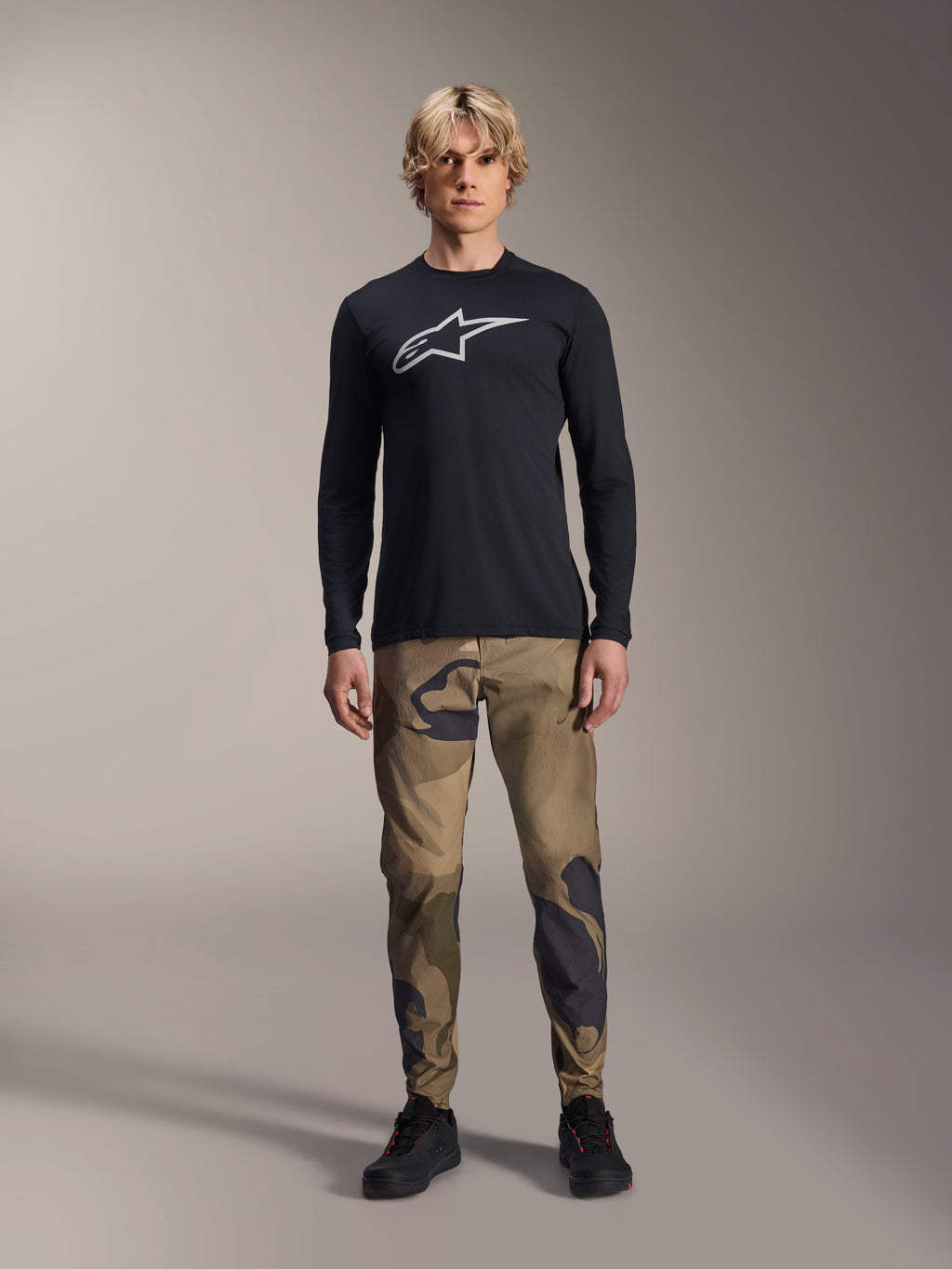 Alpinestars A-Dura Camo Pant, pantalon de cyclisme VTT, camouflage marron et vert, porté avec un maillot noir à manches longues et des chaussures noires, style VTT sportif avec tissu extensible durable