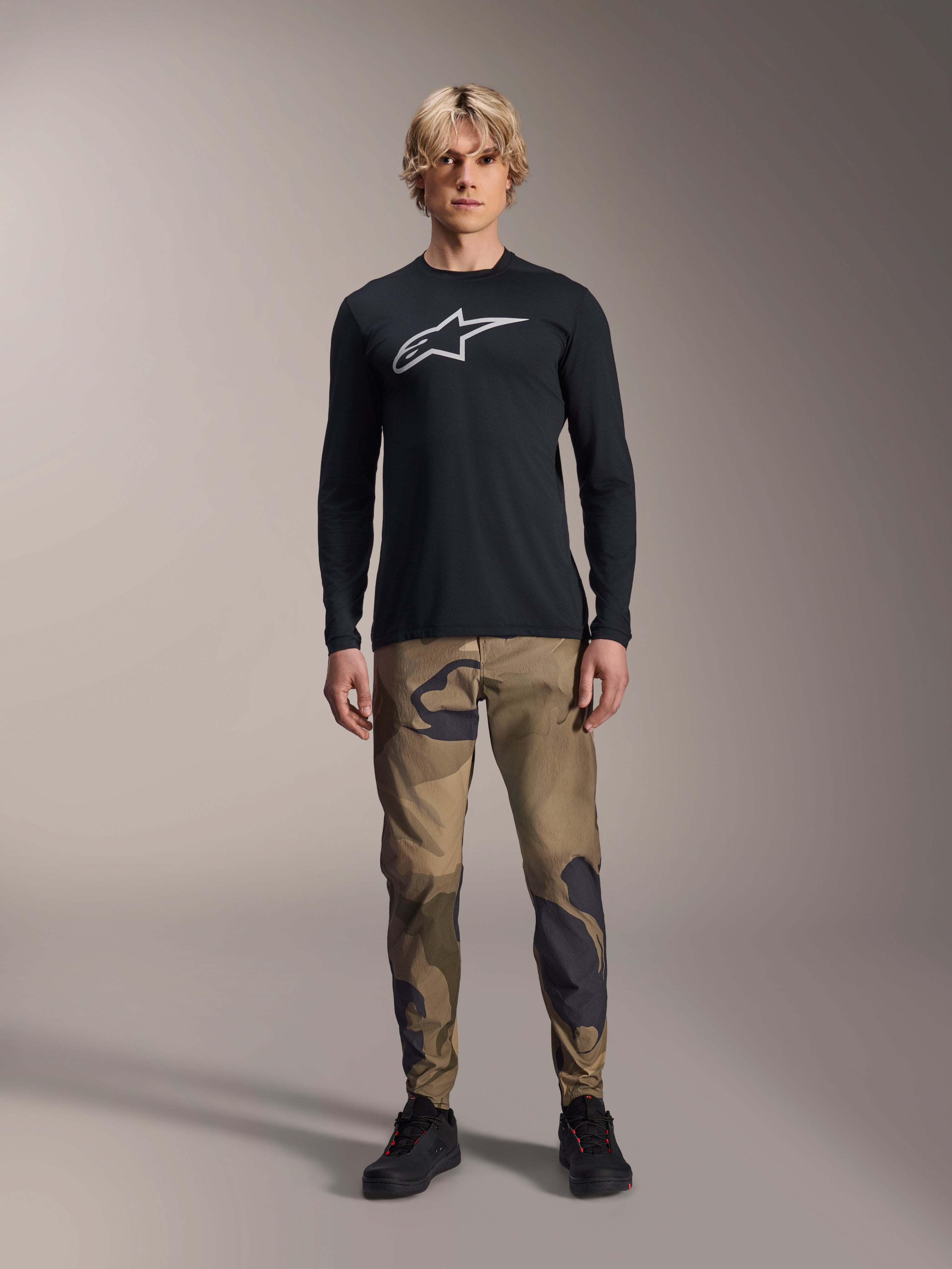 Alpinestars A-Dura Camo Pant, pantalon de cyclisme VTT, camouflage marron et vert, porté avec un maillot noir à manches longues et des chaussures noires, style VTT sportif avec tissu extensible durable