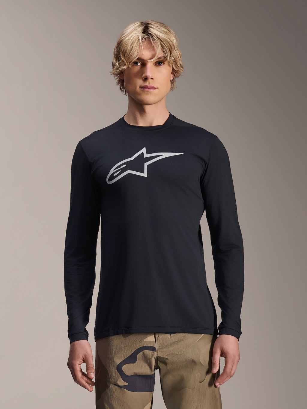 Maillot à manches longues Alpinestars A-Dura Astar, noir avec logo Astars argenté sur la poitrine, porté par un mannequin avec un pantalon camouflage, vêtement de performance VTT décontracté pour le vélo de montagne