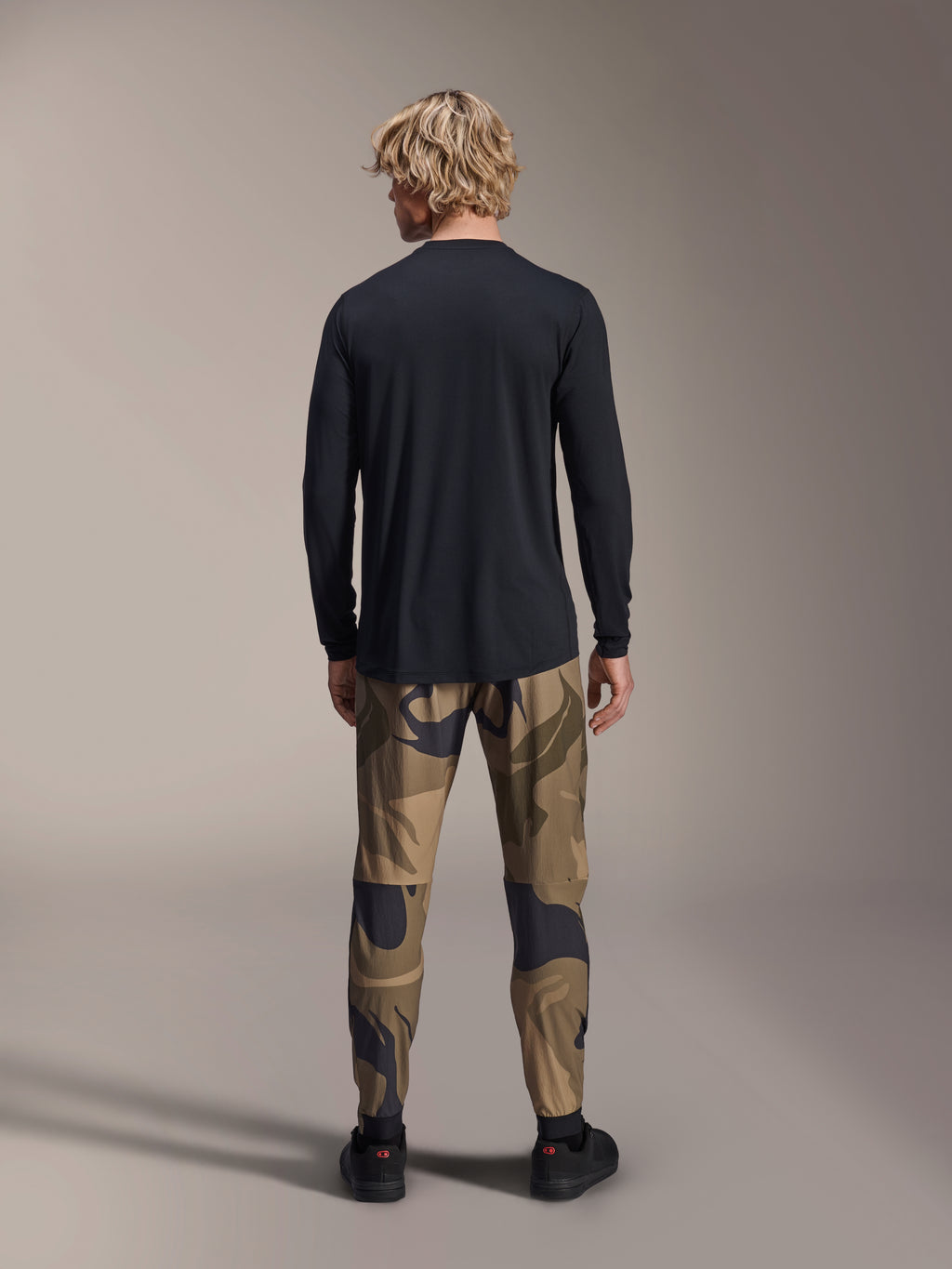 Alpinestars A-Dura Camo Pant, pantalon de VTT, motif camouflage brun, vue arrière, porté avec un maillot noir à manches longues et des baskets noires, pantalon textile à coupe régulière pour le cyclisme tout-terrain