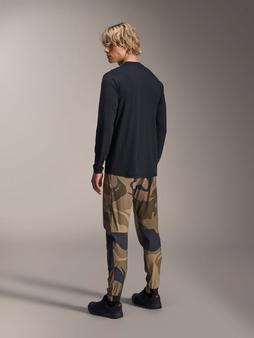 Maillot à manches longues Alpinestars A-Dura Astar, noir, vue de dos sur un mannequin, porté avec un pantalon camouflage, équipement de performance VTT décontracté avec un pan arrière plus long et une coupe régulière