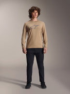 Alpinestars A-Dura Astar Jersey met lange mouwen, lichtbruin met grijs Astars-logo op de borst, gedragen door een model met een zwarte technische broek en schoenen, casual mountainbike-prestatiekleding