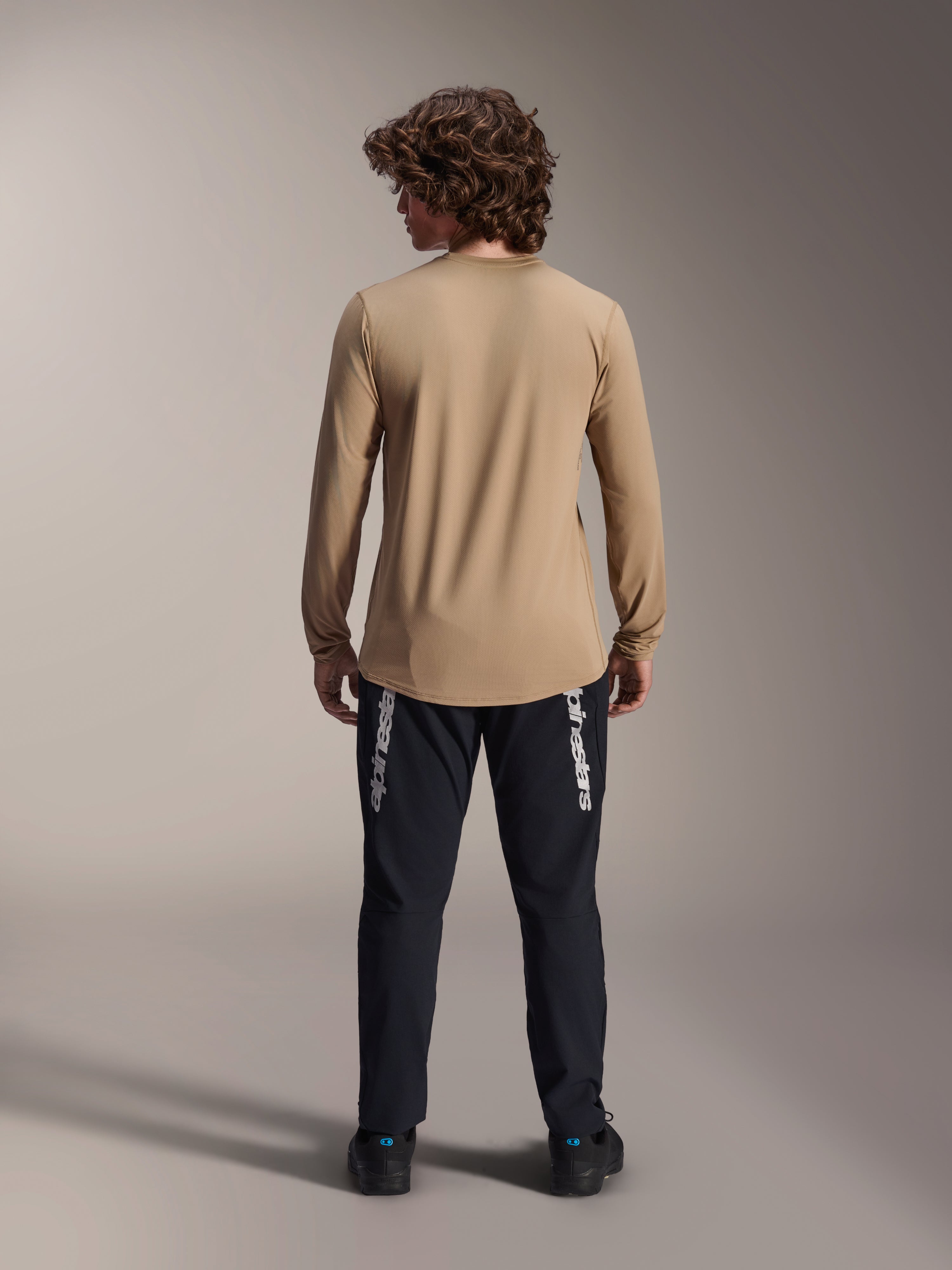 Alpinestars A-Dura Astar Jersey met lange mouwen, lichtbruin, achteraanzicht met de langere drop tail, gedragen met een zwarte broek voorzien van Alpinestars-branding, hoogwaardige mountainbike-kleding
