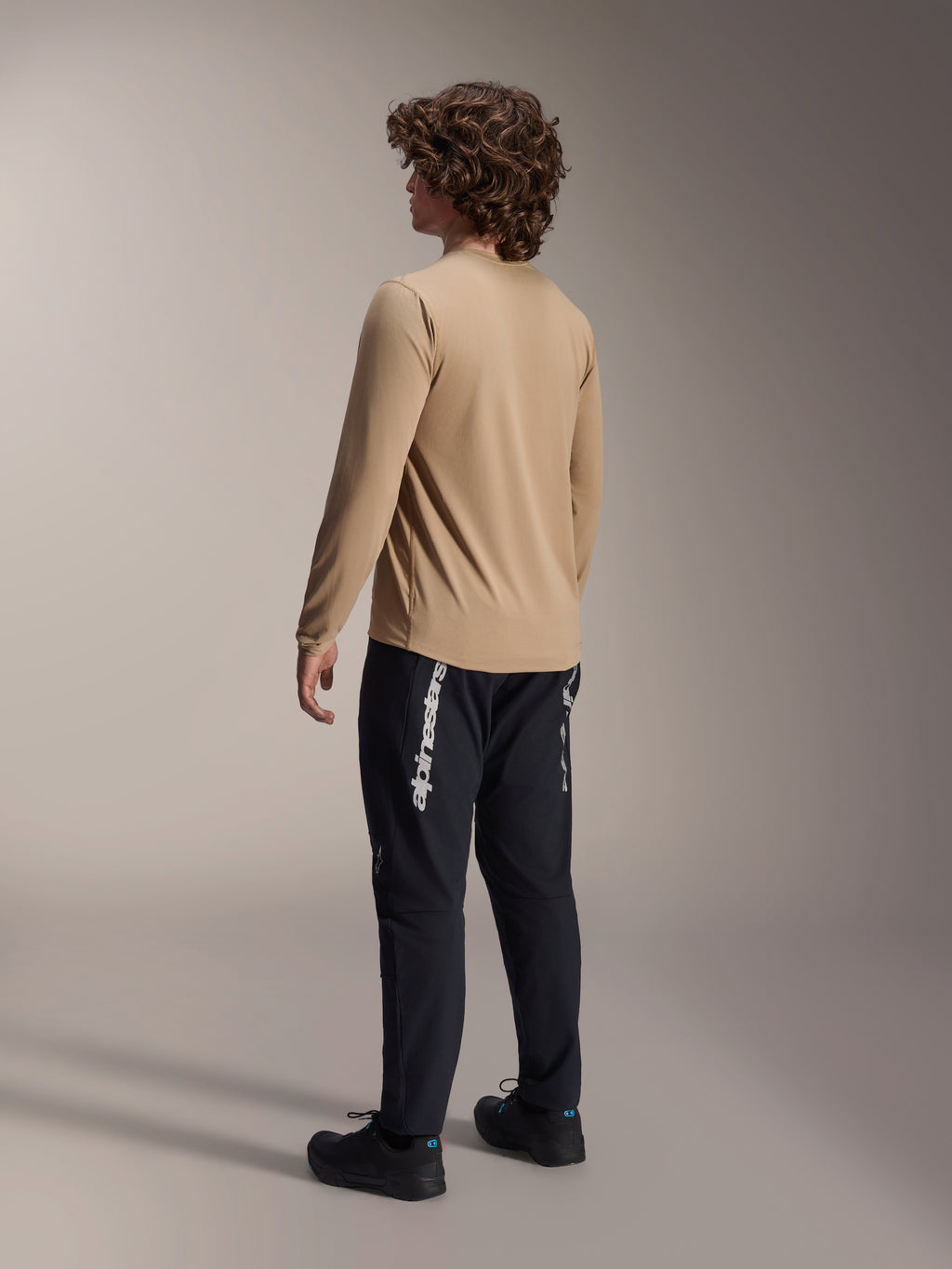 Alpinestars A-Dura Astar Long Sleeve Jersey in lichtbruin, achteraanzicht van een mannelijk model toont het drop tail-ontwerp, gecombineerd met zwarte broek en schoenen, casual mountainbike-prestatiekleding