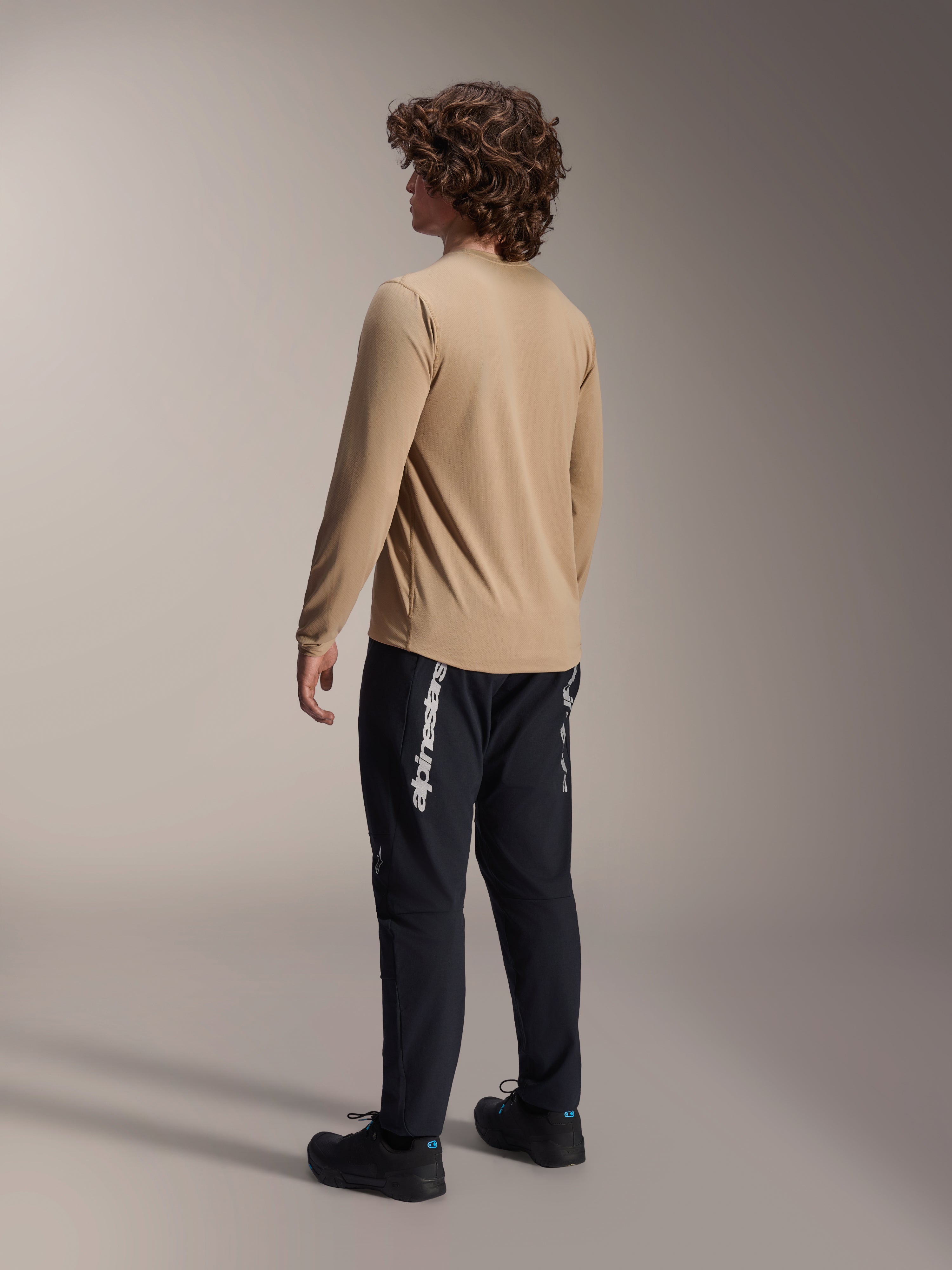 Alpinestars A-Dura Astar Long Sleeve Jersey in lichtbruin, achteraanzicht van een mannelijk model toont het drop tail-ontwerp, gecombineerd met zwarte broek en schoenen, casual mountainbike-prestatiekleding