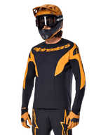Alpinestars A-Supra Race Jersey à manches longues, maillot de VTT noir et orange, porté avec casque et gants assortis, avec logo Alpinestars sur la poitrine et accents stylisés sur les manches, coupe performance pour la course tout-terrain