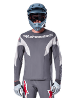 Pilote de VTT portant le maillot Alpinestars A-Supra Race - Manches longues en gris foncé, maillot de cyclisme à manches longues avec logo Alpinestars blanc, associé à un casque intégral gris-bleu, un masque et des gants de course gris, équipement de VTT complet pour la performance tout-terrain