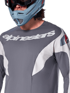 Alpinestars A-Supra Race Jersey - Manches longues, maillot de VTT, gris foncé avec graphismes de course blancs et logo Alpinestars blanc, porté avec un casque intégral bleu clair et des lunettes, tissu micro-perforé pour la performance tout-terrain