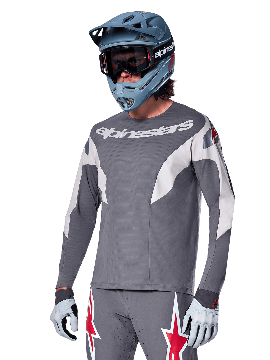 Maillot Alpinestars A-Supra Race à manches longues, gris foncé avec accents blancs et rouges, graphismes de style course, porté avec un pantalon gris assorti, un casque intégral bleu et des gants blancs, équipement professionnel de VTT et de descente