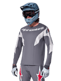 Maillot Alpinestars A-Supra Race à manches longues, gris foncé avec accents blancs et rouges, graphismes de style course, porté avec un pantalon gris assorti, un casque intégral bleu et des gants blancs, équipement professionnel de VTT et de descente