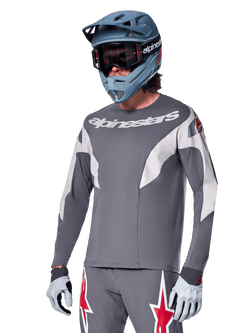 Alpinestars A-Supra Race Trikot – Langarm, Dunkelgrau mit weißen und roten Akzenten, Grafiken im Rennstil, kombiniert mit passender grauer Hose, blauem Fullface-Helm und weißen Handschuhen, professionelle MTB- und Downhill-Rennausrüstung