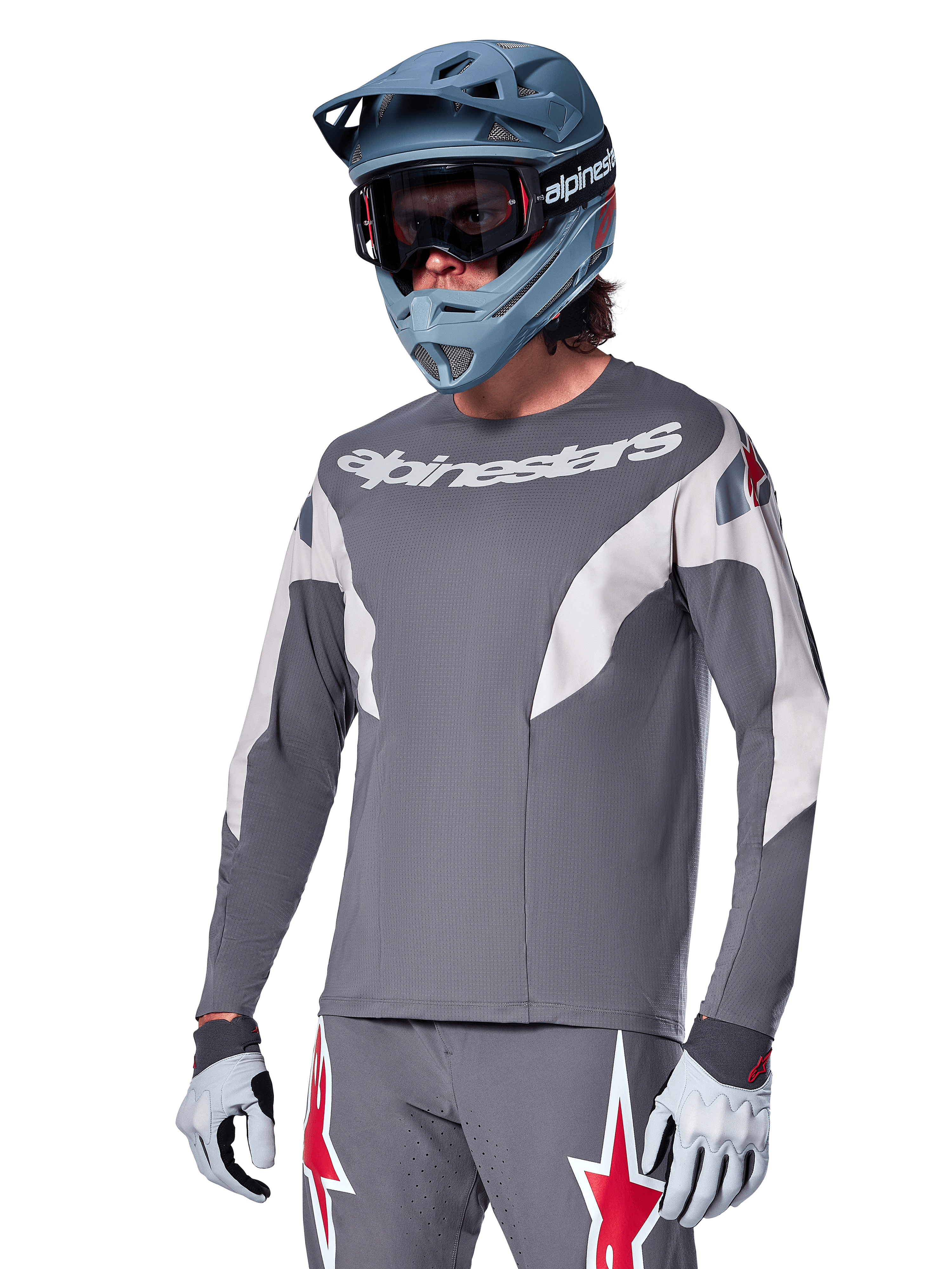 Maillot Alpinestars A-Supra Race à manches longues, gris foncé avec accents blancs et rouges, graphismes de style course, porté avec un pantalon gris assorti, un casque intégral bleu et des gants blancs, équipement professionnel de VTT et de descente