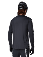 Maillot à manches longues Alpinestars A-Dura Switch, noir, vue arrière sur un cycliste portant un casque de VTT noir et des gants, doté d'un tissu jacquard technique et d'une coupe allongée dans le dos pour la pratique du tout-terrain