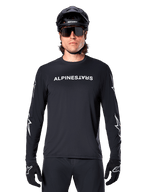 Maillot à manches longues Alpinestars A-Dura Switch, maillot de VTT noir, logo Alpinestars blanc sur la poitrine et graphismes d'étoiles sur les manches, porté avec un casque de VTT noir et des lunettes de soleil de performance, vêtement technique de trail