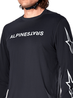 Maillot à manches longues Alpinestars A-Dura Switch, noir, avec logo Alpinestars blanc sur la poitrine et logos étoiles sur la manche, porté par un cycliste avec une sangle de casque visible, vêtement technique de VTT