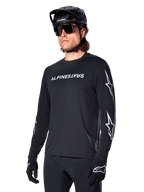 Cycliste masculin portant le maillot à manches longues Alpinestars A-Dura Switch, noir avec marquage blanc et logos étoiles sur les manches, avec casque de VTT noir et lunettes de soleil, équipement de cyclisme haute performance pour le tout-terrain