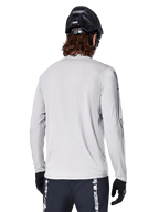 Maillot à manches longues Alpinestars A-Dura Switch, gris clair, vue arrière d'un vététiste portant un casque de VTT noir et des gants, avec une coupe arrière plus longue pour une protection accrue et un tissu jacquard texturé, équipement de cyclisme haute performance