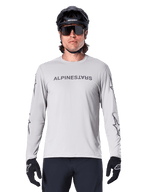 Maillot à manches longues Alpinestars A-Dura Switch, gris clair, avec logo textuel gris foncé sur la poitrine et logos étoiles sur les manches, porté par un cycliste avec un casque de VTT noir et des gants, vêtement technique performant pour le trail