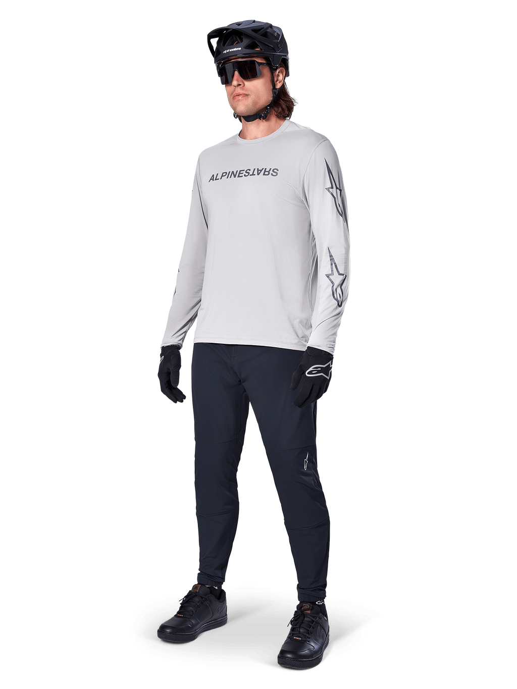 Maillot à manches longues Alpinestars A-Dura Switch, gris clair, porté par un cycliste avec un pantalon de VTT noir et un casque, arborant un logo lettrage noir sur la poitrine et des graphismes d'étoiles sur les manches, équipement de cyclisme tout-terrain haute performance