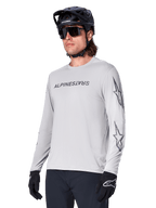 Maillot à manches longues Alpinestars A-Dura Switch, maillot de VTT, gris clair, inscription noire sur la poitrine et logos étoiles sur les manches, porté avec un casque de VTT noir, des lunettes de soleil sombres et des gants noirs, équipement de cyclisme tout-terrain athlétique