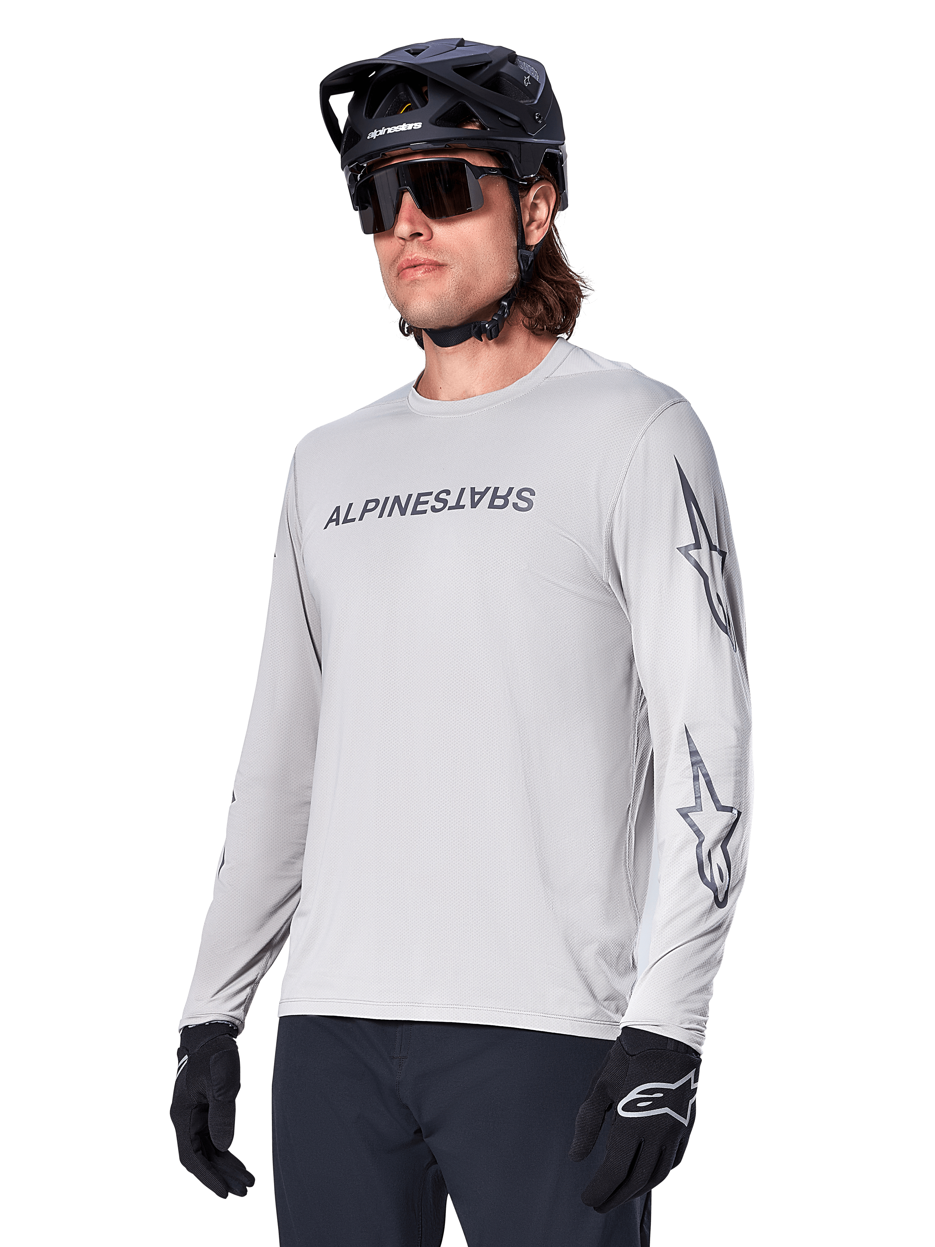 Maillot à manches longues Alpinestars A-Dura Switch, maillot de VTT, gris clair, inscription noire sur la poitrine et logos étoiles sur les manches, porté avec un casque de VTT noir, des lunettes de soleil sombres et des gants noirs, équipement de cyclisme tout-terrain athlétique