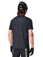 Alpinestars A-Dura Switch Jersey met korte mouwen, zwart, achteraanzicht op een model met een zwarte fietshelm en MTB-short, normale pasvorm en langere achterzijde voor mountainbiken