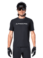 Mountainbiker die het Alpinestars A-Dura Switch Jersey met korte mouwen in het zwart draagt, met een wit woordmerklogo op de borst, gecombineerd met een zwarte helm, grote zonnebril en zwarte handschoenen, professionele fietskleding voor trailrijden.