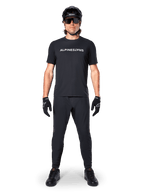 Mountainbiker die het Alpinestars A-Dura Switch Jersey met korte mouwen in het zwart draagt, voorzien van een wit gestileerd woordmerk-logo op de borst, gecombineerd met bijpassende zwarte MTB-broek, helm en handschoenen, complete trail-ready wieleroutfit.