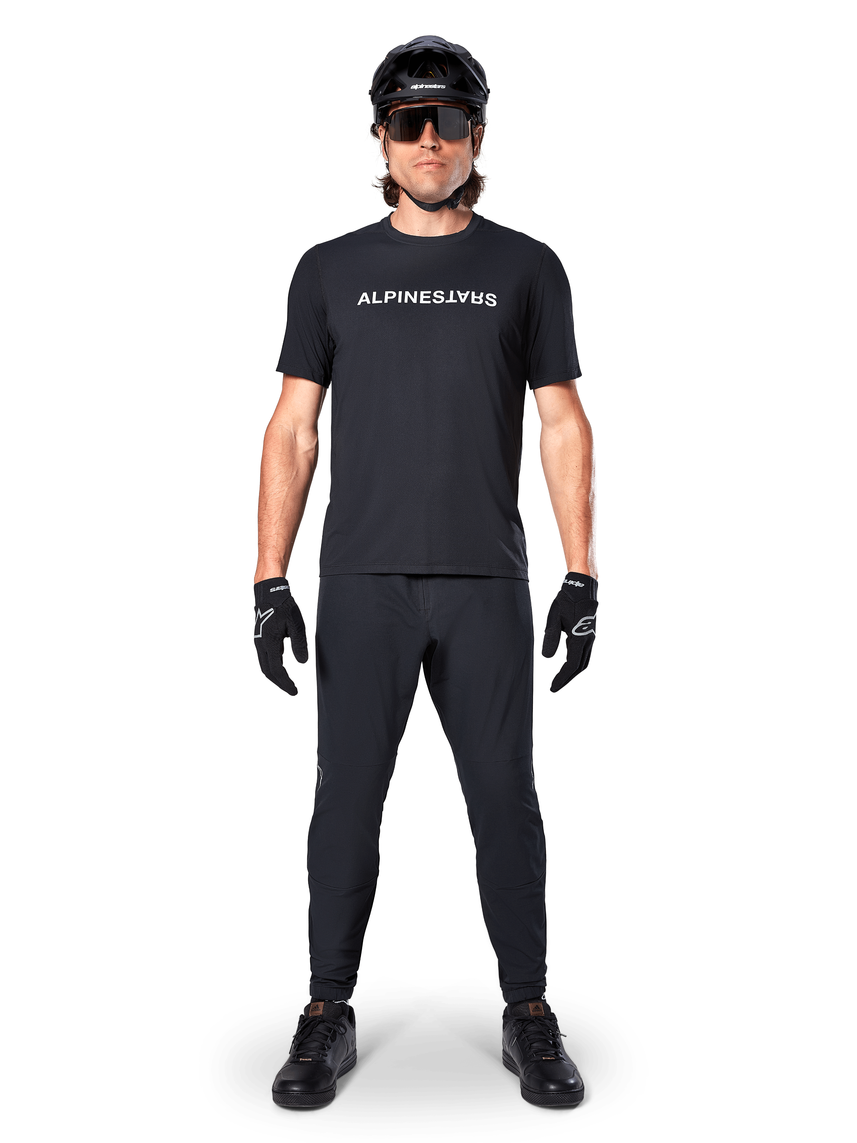 Mountainbiker die het Alpinestars A-Dura Switch Jersey met korte mouwen in het zwart draagt, voorzien van een wit gestileerd woordmerk-logo op de borst, gecombineerd met bijpassende zwarte MTB-broek, helm en handschoenen, complete trail-ready wieleroutfit.