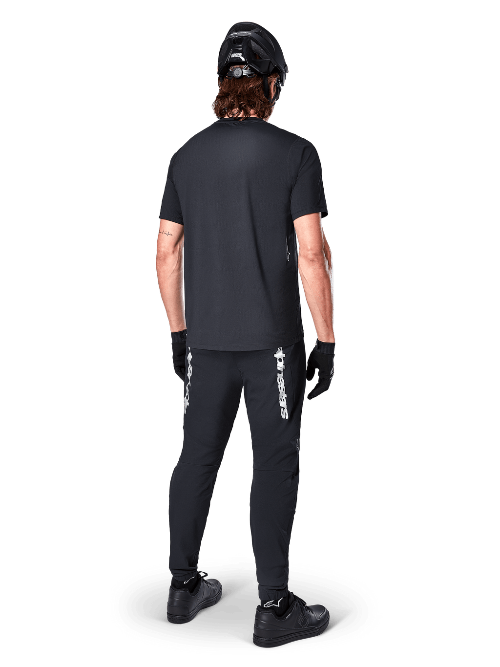Alpinestars A-Dura Switch Jersey - Korte Mouwen, zwart, achteraanzicht van rijder in volledige mountainbike-uitrusting, inclusief bijpassende zwarte broek met wit woordmerk-logo, zwarte helm en handschoenen, technische fietskleding voor trailrijden