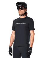 Alpinestars A-Dura Switch jersey met korte mouwen, zwart, wit woordmerklogo op de borst, gedragen door een fietser met zwarte MTB-helm en zonnebril, technische kleding voor trailrijden