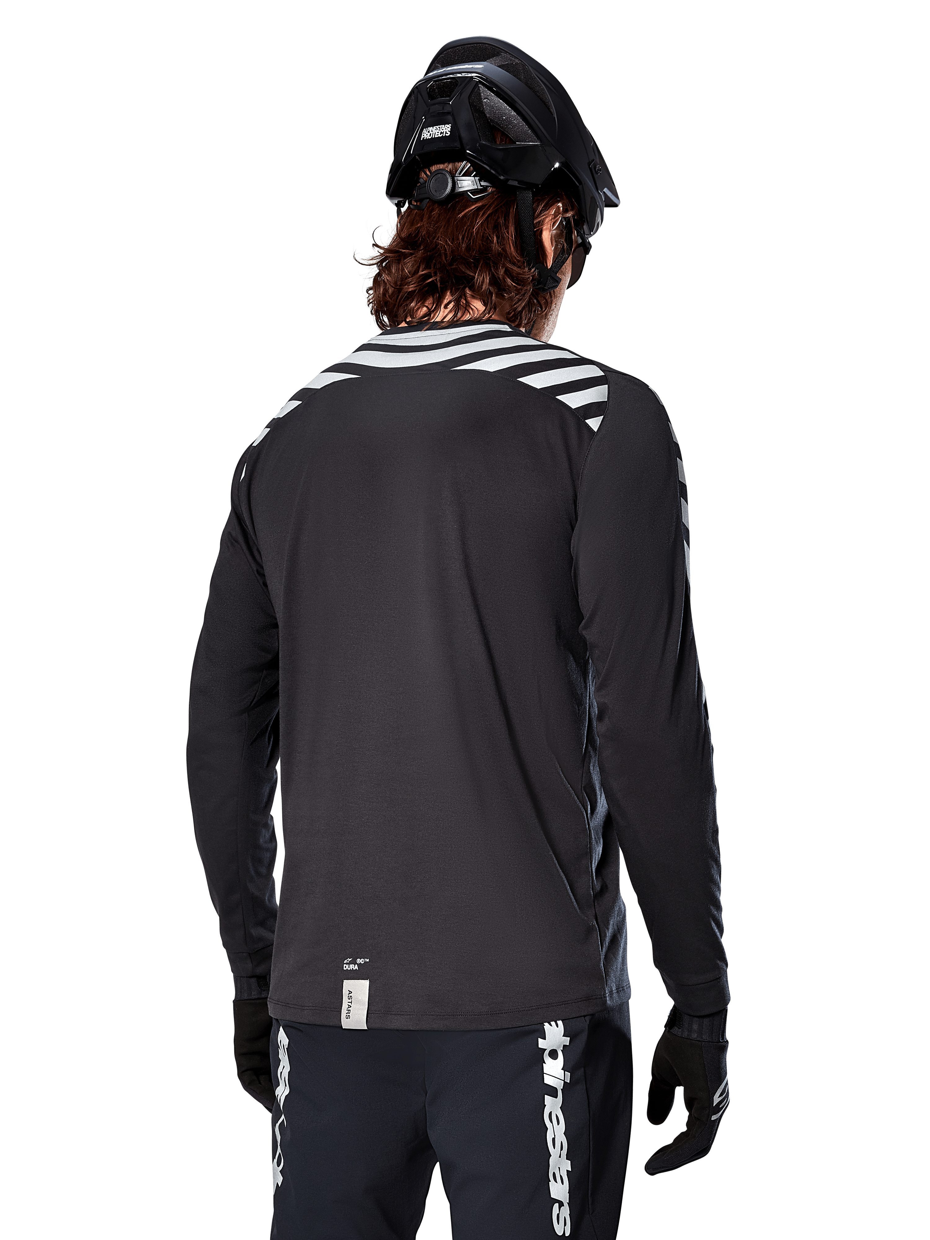 Alpinestars A-Dura Dri Oscar Long Sleeve Jersey, Black Reflective, rear view showing reflective diagonal stripes on shoulders and a longer drop tail, worn with black MTB helmet and gloves, technical gear for mountain biking