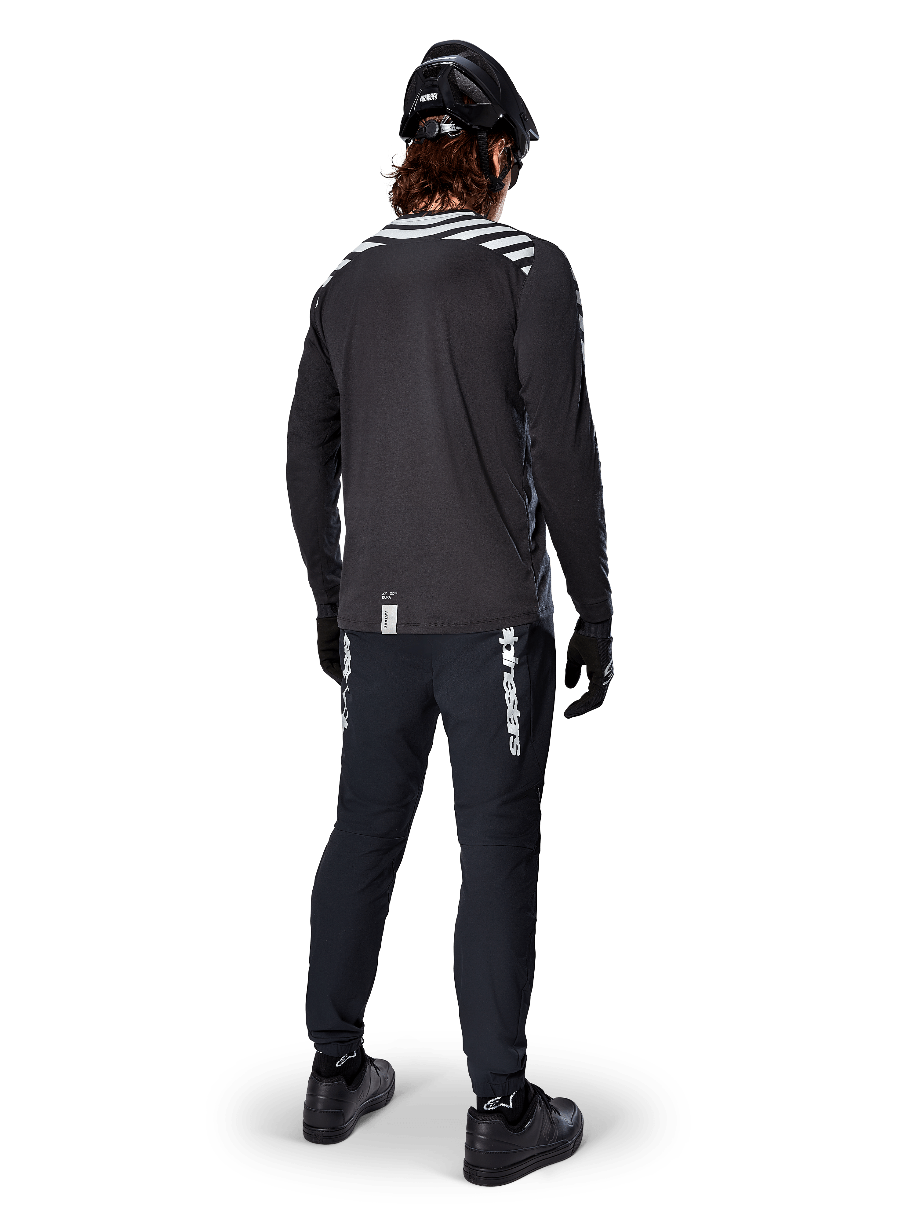 Mountain bike rider wearing the Alpinestars A-Dura Dri Oscar Long Sleeve Jersey in Black Reflective, viewed from behind, paired with black technical pants, helmet, and gloves, featuring reflective stripe graphics on the shoulders and a longer drop tail for off-road cycling.