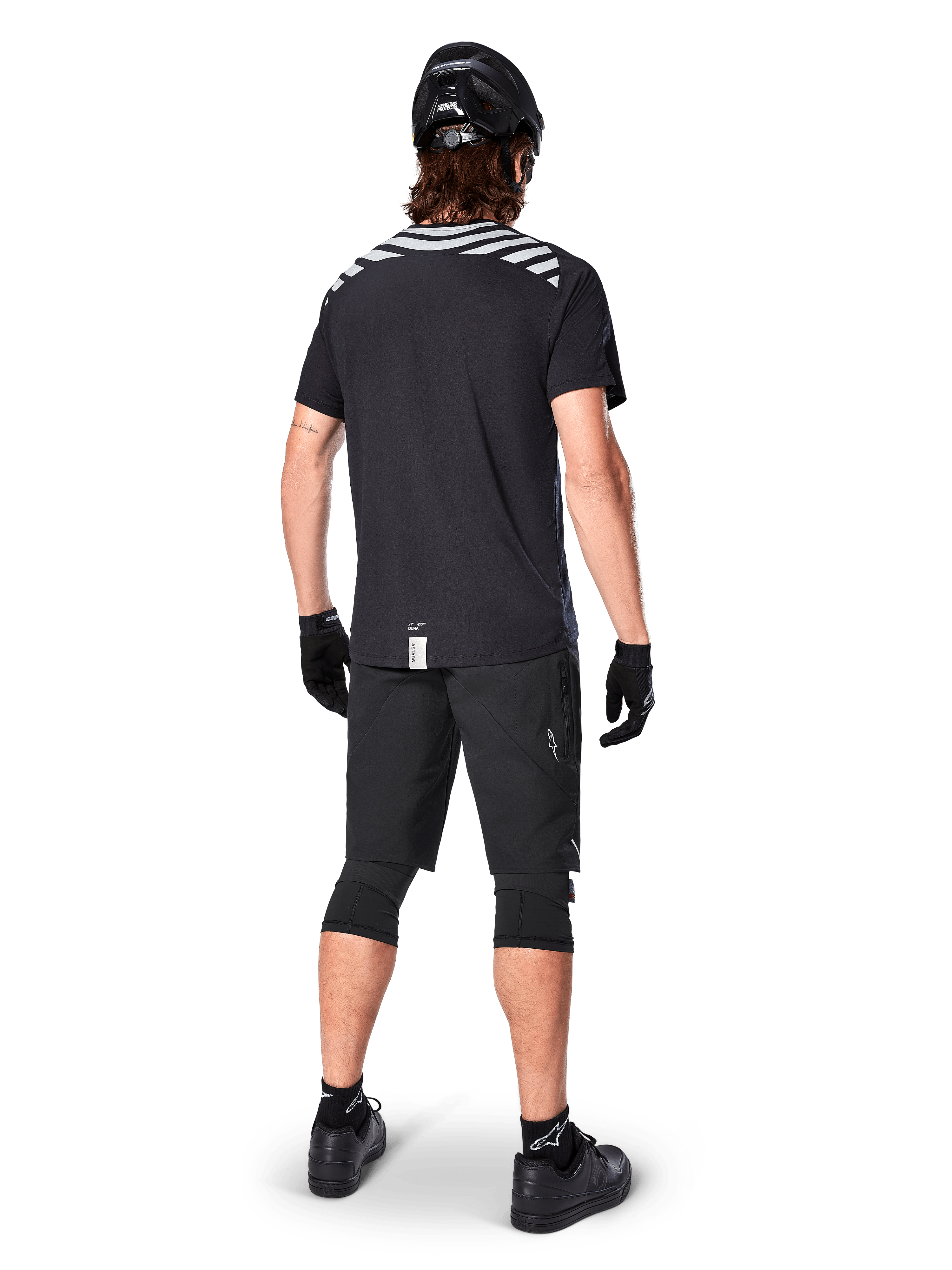 Alpinestars A-Dura Dri Oscar Kurzarmtrikot, Black Reflective, Rückansicht eines Fahrers mit schwarzen Mountainbike-Shorts, Helm und Knieschonern, reflektierende Schultergrafiken und verlängerte Rückenpartie für MTB-Fahrten.