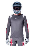 MTB-fietser in Alpinestars A-Supra Astar Jersey met lange mouwen, donkergrijs met rode graphics op de mouwen en witte schouderaccenten, bijpassende grijze broek met rode ster-logo's, blauwe full-face helm en goggle, gecoördineerde off-road wieleruitrusting