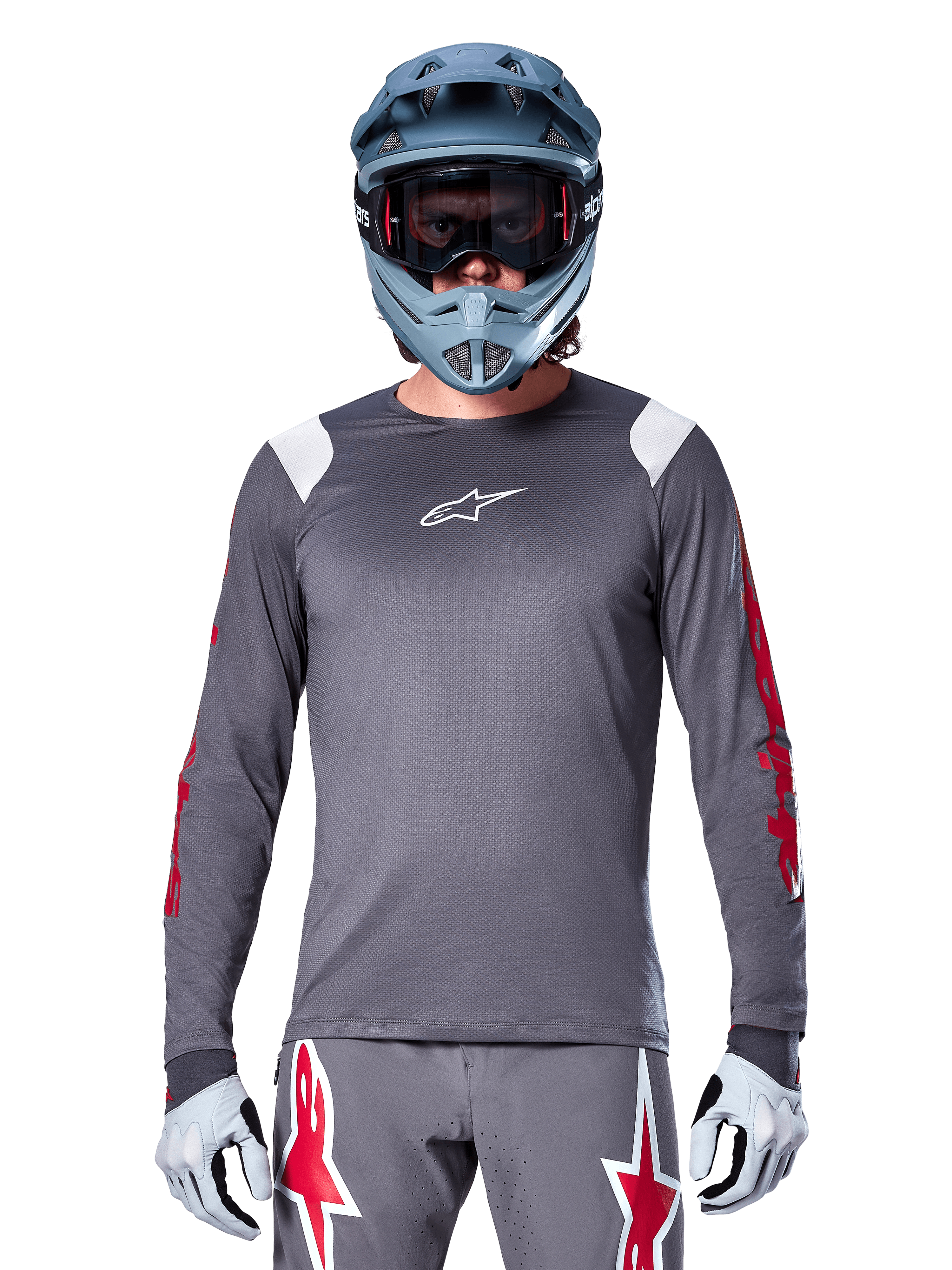 MTB-fietser in Alpinestars A-Supra Astar Jersey met lange mouwen, donkergrijs met rode graphics op de mouwen en witte schouderaccenten, bijpassende grijze broek met rode ster-logo's, blauwe full-face helm en goggle, gecoördineerde off-road wieleruitrusting