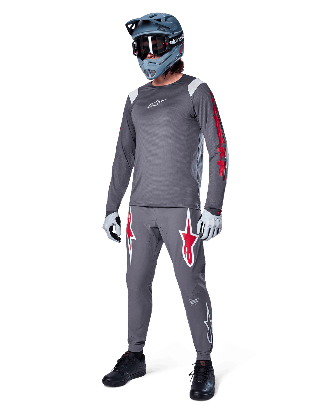 Alpinestars A-Supra Astar Jersey met lange mouwen, donkergrijs met rode branding op de mouwen en wit logo op de borst, gedragen door een rijder met bijpassende grijze broek, blauwgrijze full-face helm en handschoenen, complete mountainbike-uitrusting voor gravity riding.
