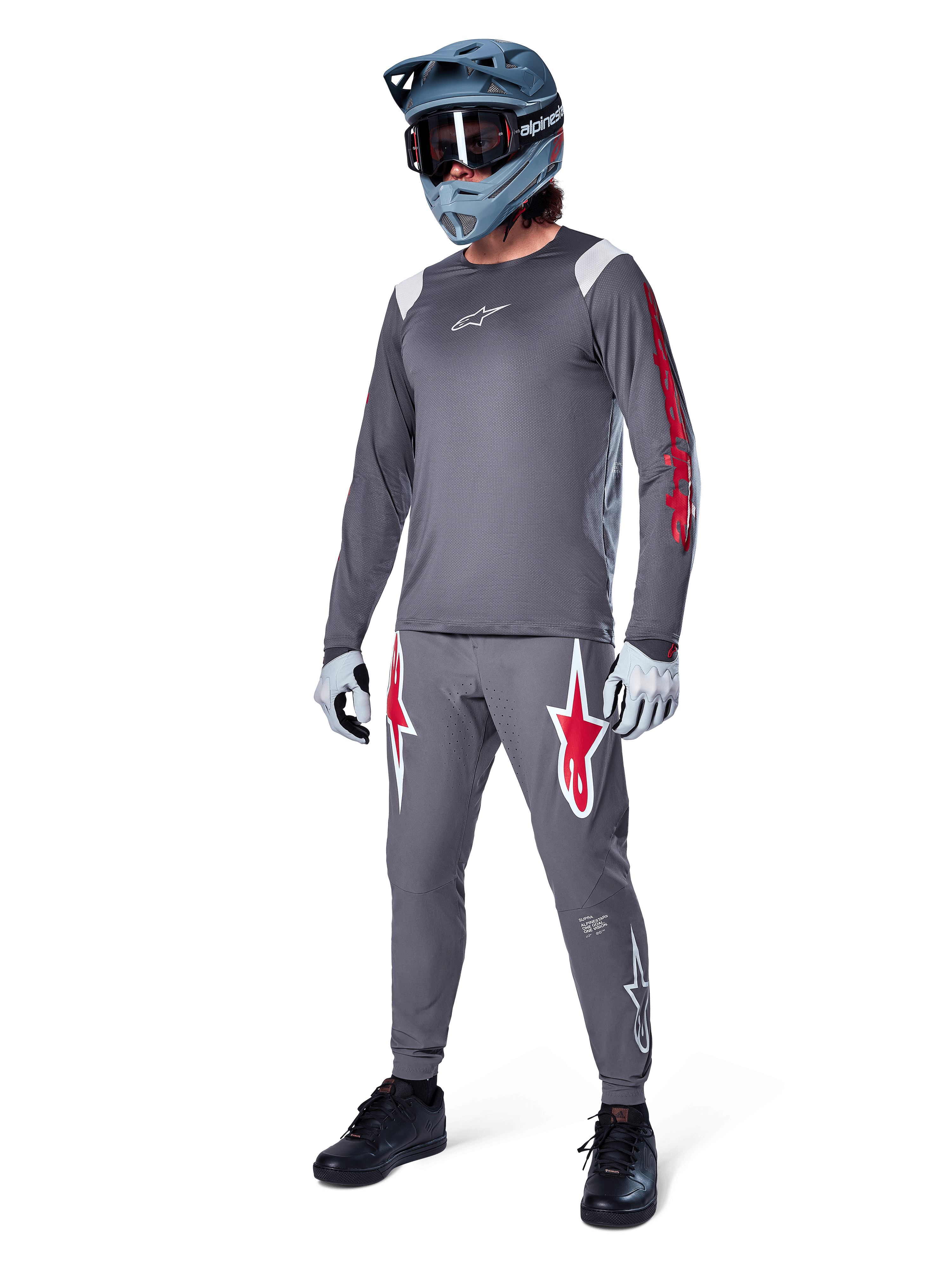 Alpinestars A-Supra Astar Jersey met lange mouwen, donkergrijs met rode branding op de mouwen en wit logo op de borst, gedragen door een rijder met bijpassende grijze broek, blauwgrijze full-face helm en handschoenen, complete mountainbike-uitrusting voor gravity riding.
