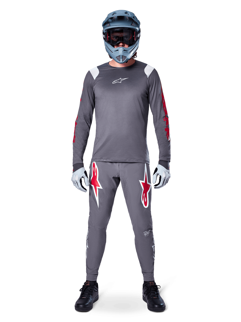 MTB-rijder in Alpinestars A-Supra Astar Jersey met lange mouwen, donkergrijs met rode accenten en wit logo op de borst, bijpassende grijze broek met grote rode ster-graphics, blauwe full-face helm met crossbril, lichtgrijze handschoenen en zwarte schoenen, professionele mountainbike-uitrusting