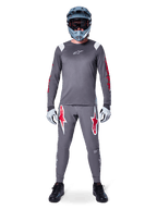 MTB-rijder in Alpinestars A-Supra Astar Jersey met lange mouwen, donkergrijs met rode accenten en wit logo op de borst, bijpassende grijze broek met grote rode ster-graphics, blauwe full-face helm met crossbril, lichtgrijze handschoenen en zwarte schoenen, professionele mountainbike-uitrusting