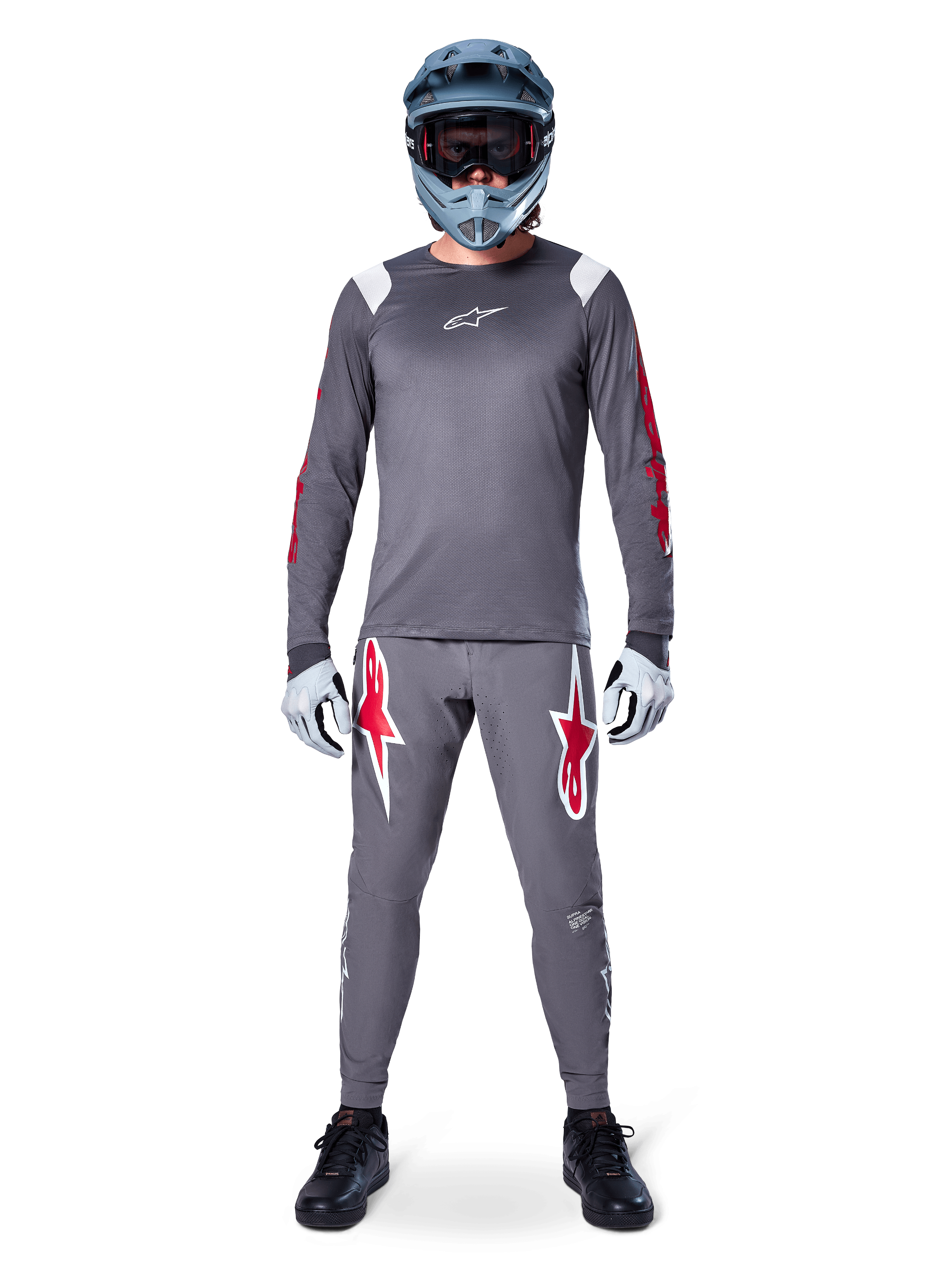 MTB-rijder in Alpinestars A-Supra Astar Jersey met lange mouwen, donkergrijs met rode accenten en wit logo op de borst, bijpassende grijze broek met grote rode ster-graphics, blauwe full-face helm met crossbril, lichtgrijze handschoenen en zwarte schoenen, professionele mountainbike-uitrusting