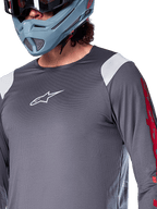 Alpinestars A-Supra Astar Jersey met lange mouwen, donkergrijs met witte schouderpanelen en rode graphics op de mouwen, gedragen met een licht blauwgrijze full-face helm, performance mountainbikekleding